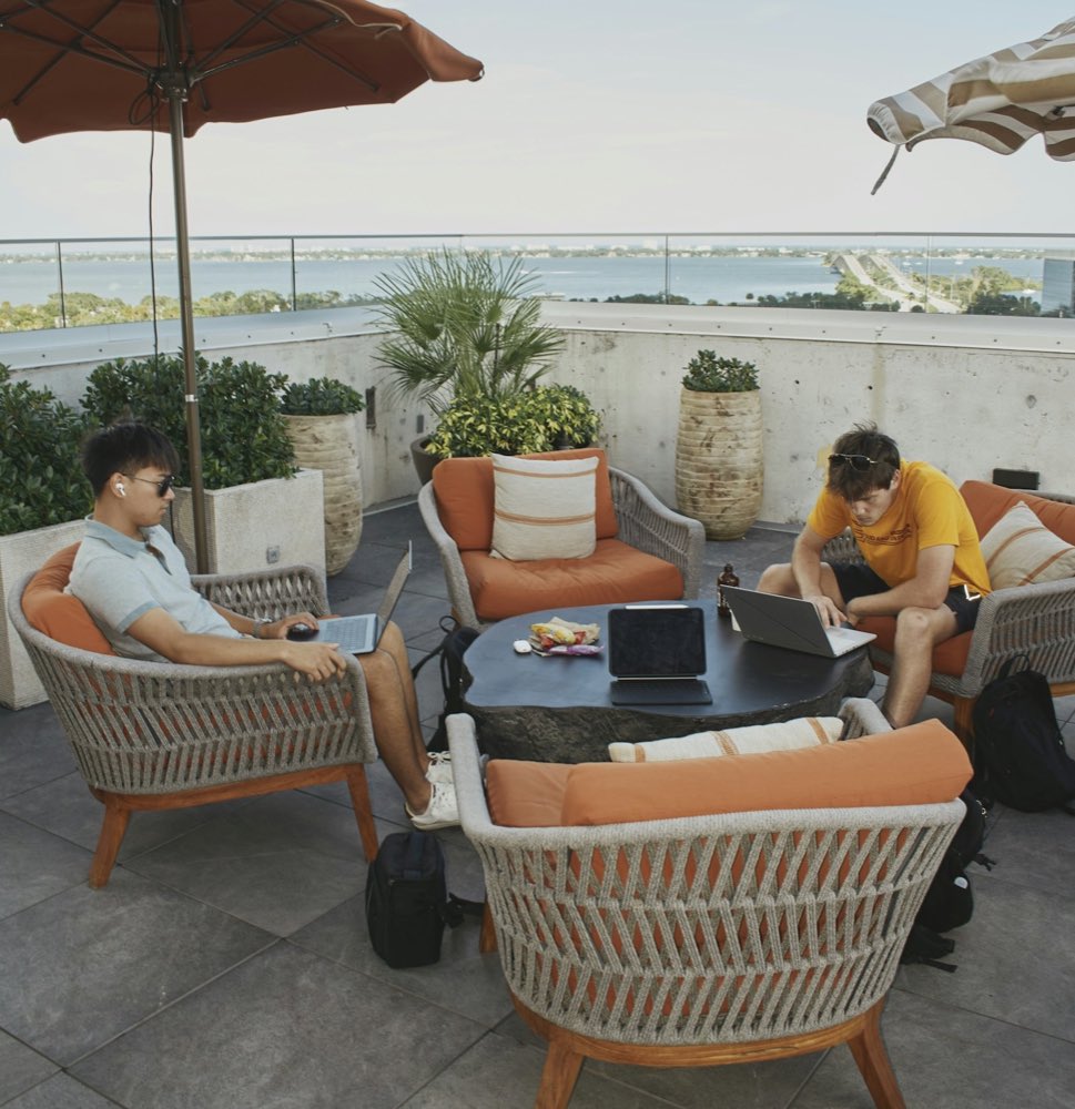 Two people working on laptops on a rooftop terrace with a sea view, under a terracotta parasol